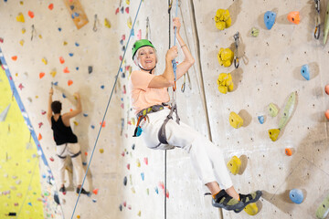 Confident senior woman wearing helmet scaling artificial climbing wall in climbing gym
