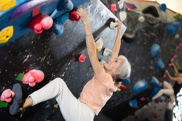 Fototapeta na wymiar Sporty elderly female exercising in boulder climbing hall without rope, reaching new results, enjoying new challenges in amusement park