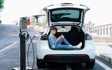 Fototapeta na wymiar Little boy sitting on car trunk, using smartphone while recharging eco-friendly car from EV charging station. EV car road trip travel as alternative vehicle using sustainable energy concept. Perpetual