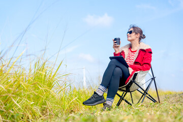 Beautiful Caucasian woman sit on chair near meadow with blue sky and wind turbines or windmill on the back and she also hold a cup of coffee with relax action.