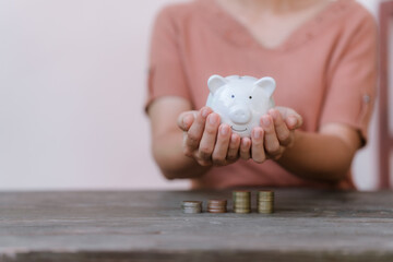 hand placing a coin into a white piggy bank on a table against a pale pink background.