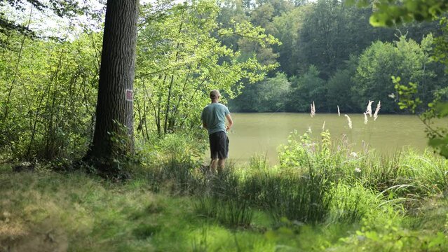 Male angler stands on shore of cloudy lake between green grass and trees catching fish with fishing rod, pan