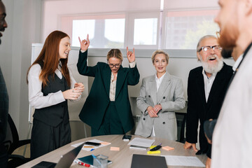 Wide shot of diverse multiracial team of happy excited business people standing around office table, celebrating launch of start up project, having fun, cheering, raising hands and laughing.
