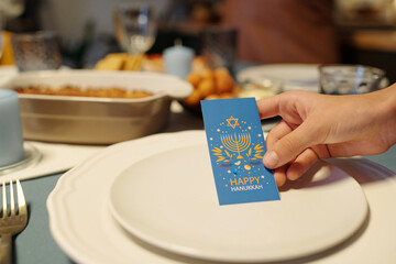 Hand of girl holding blue Hanukkah greeting card with symbols of traditional Jewish holiday over plate while serving table for dinner