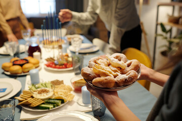 Close-up of unrecognizable girl holding plate with appetizing homemade donuts while carrying it to table served for Hanukkah dinner