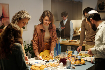 Happy Jewish family putting plates with homemade food on table while serving it for invited guests before Hanukkah dinner