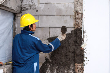 Asian male house builder with yellow helmet and uniform applying cement on an outdoor wall with...