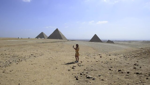 Influencer girl telecommuting records video with her mobile and talks to camera in front of egyptian pyramids in Gyza, Egypt