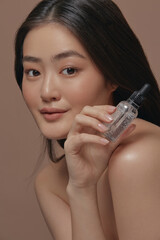 Vertical close up portrait, beautiful korean ethnicity woman with a healthy clean and perfectly smooth skin holding facial serum near self face posing over beige isolated background. 