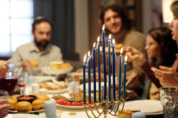 Burning candles on menorah candlestick standing on served table with homemade food and drinks against members of Jewish family