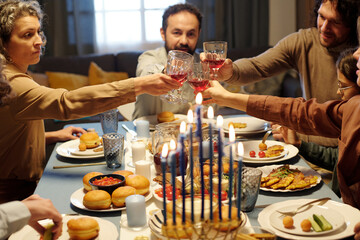 Members of large Jewish family clinking with wineglasses over served table with homemade food while...