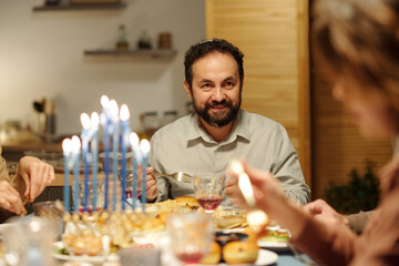 Smiling Jewish man looking at his wife or other family member during Hanukkah dinner while having...