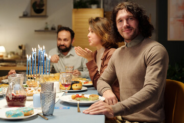 Happy young Jewish man looking at camera while sitting by table served with homemade food and drinks against his wife and father