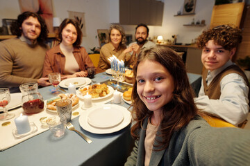 Cute smiling girl taking selfie with members of her family by dinner while sitting by served table with homemade food and drinks