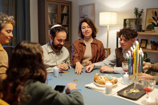 Young woman sliding coin on table while playing leisure game with members of her family sitting by table with menorah candlestick