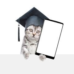 Сollege graduate kitten holds big smartphone with white blank screen in it paw, showing above empty white banner. isolated on white background