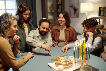 Young cheerful woman laughing while sitting next to mature man spinning small dreidel during...