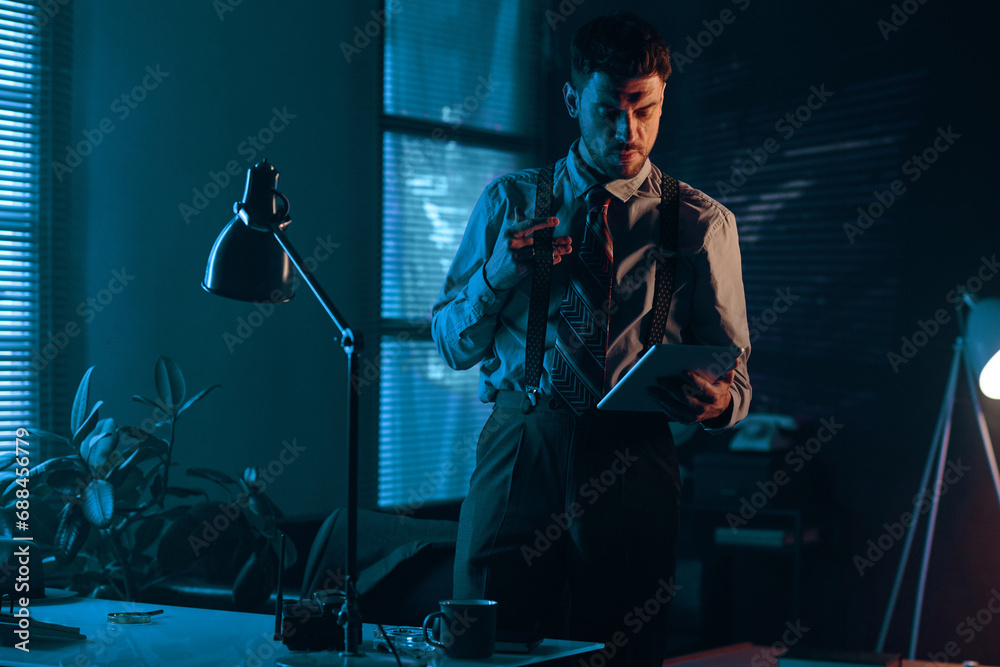 Poster young man in retro formalwear looking at screen of tablet while standing by his workplace in dark of