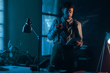 Young man in retro formalwear looking at screen of tablet while standing by his workplace in dark office and scrolling through online data