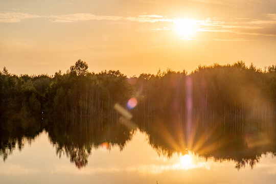This image embodies the ethereal beauty of a sunset reflected on a lake, with the sun descending towards the horizon amidst a soft golden haze. The calm surface of the water creates a perfect mirror
