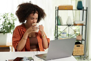 Woman with fluffy curly hair drinking cup of coffee and watching product presentation or...