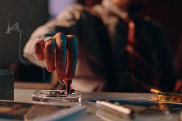 Close-up of hand of young man stubbing butt of cigarette in ashtray standing on table among business and office supplies