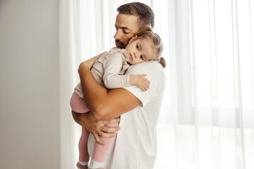 Caring dad is holding and hugging his daughter while standing at home.