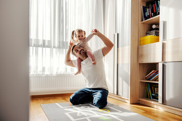 Playful dad is holding his daughter on his shoulders and playing in nursery room.