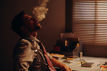 Fototapeta premium Young relaxed man with cigarette in mouth blowing smoke while sitting by workplace in office and having break after overtime work