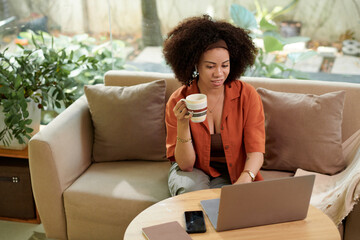 Smiling woman drinking morning coffee and reading report on her company performance when working...