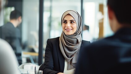 A young Muslim woman in a hijab sits at a table in an office, communicating with an interlocutor.