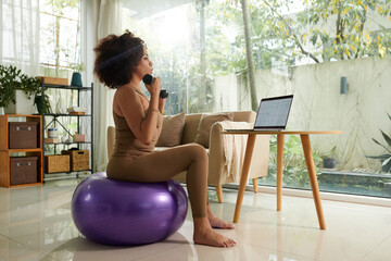 Woman exercising with dumbbells on fitness ball when checking her calendar on laptop