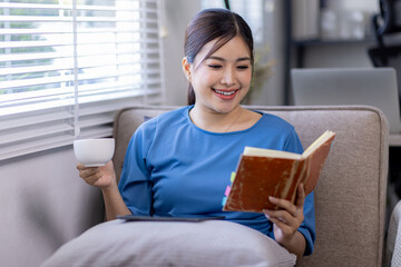 Happy asian woman sitting on a couch at home reading a paper book

