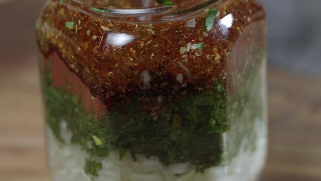 Glass jar with ingredients for chimichurri, fresh parsley, oregano, ground red pepper, black pepper, salt, garlic and more. The bubbling oil is incorporated into the mixture