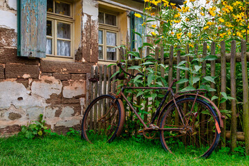 Old abandoned rusty bicycle leaning at a wooden fence in front of an old farm house