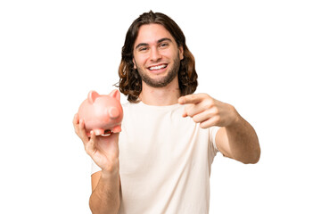 Young handsome man holding a piggybank over isolated background points finger at you with a...