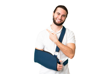 Young handsome man with broken arm and wearing a sling over isolated chroma key background pointing...
