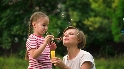 Happy mother with cheerful daughter blows bubbles in park on meadow on vacation