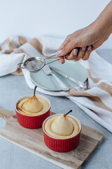 A pear soufflé in a red bowl stands on a wooden board, next to a vintage fork with a knife and a kitchen towel. A woman's hand holds a sieve and sprinkles cinnamon