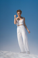 Woman walks through snow with light fog on sunny weather with blue sky. Romantic blond adult female looking down. Tourist wearing in white crop top, white pants and sandals. Full length, front view