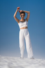 Obraz premium Sexual blond adult woman dressed in white crop top, white pants and sandals. European female tourist looking down, hands up, walking through snowfield with light fog on sunny day with blue sky