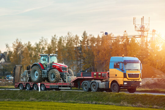 The semi truck is hauling farm equipment on a flatbed. The heavy industrial truck semi trailer flatbed platform transport one big modern farming tractor machine on the road. Evening sun.