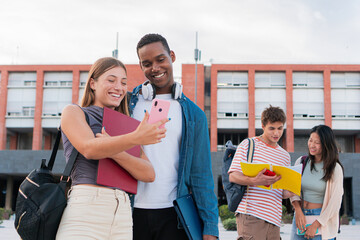 Happy young smiling college students watching something on smart phone, funny news while taking a break before starting lessons. Multiethnic couple sharing content with a cell phone inside the campus