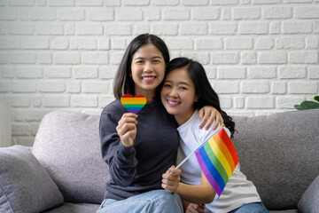 Attractive Asian lesbian couple holding LGBT flag in living room at home with smiling faces
