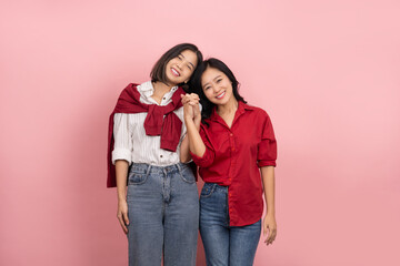 Cheerful young Asian lesbian couple in shirts and jeans stands holding hands to express their love...