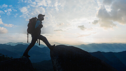 Low angle view of Asian male hikers climbing mountain at sunset rays over the clouds with trekking poles on cliff edge on top of rock mountain, Successful summit concept image.