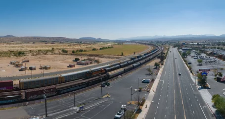 Poster Aerial view of a serene semi-arid landscape with mountains, a road with parked vehicles, a moving freight train, and a modern town under a clear blue sky. © ingusk
