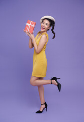 happy smiling fun asian girl in yellow dress holding present box raise up leg isolated over purple background.