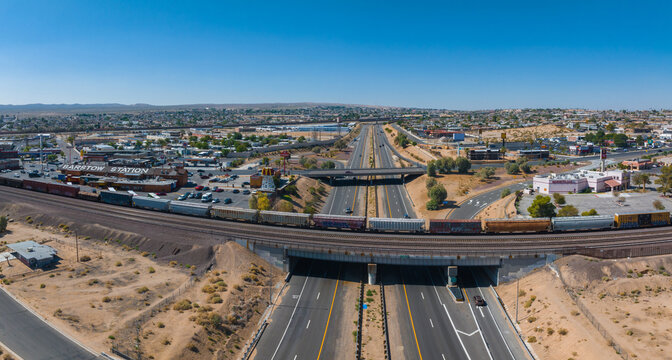 Aerial view of a southwestern American town with a freight train crossing a highway, surrounded by desert terrain, commercial and residential buildings under a clear blue sky.