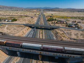 Aerial view of a sunlit highway interchange and parallel train tracks in a desert landscape. Sparse...